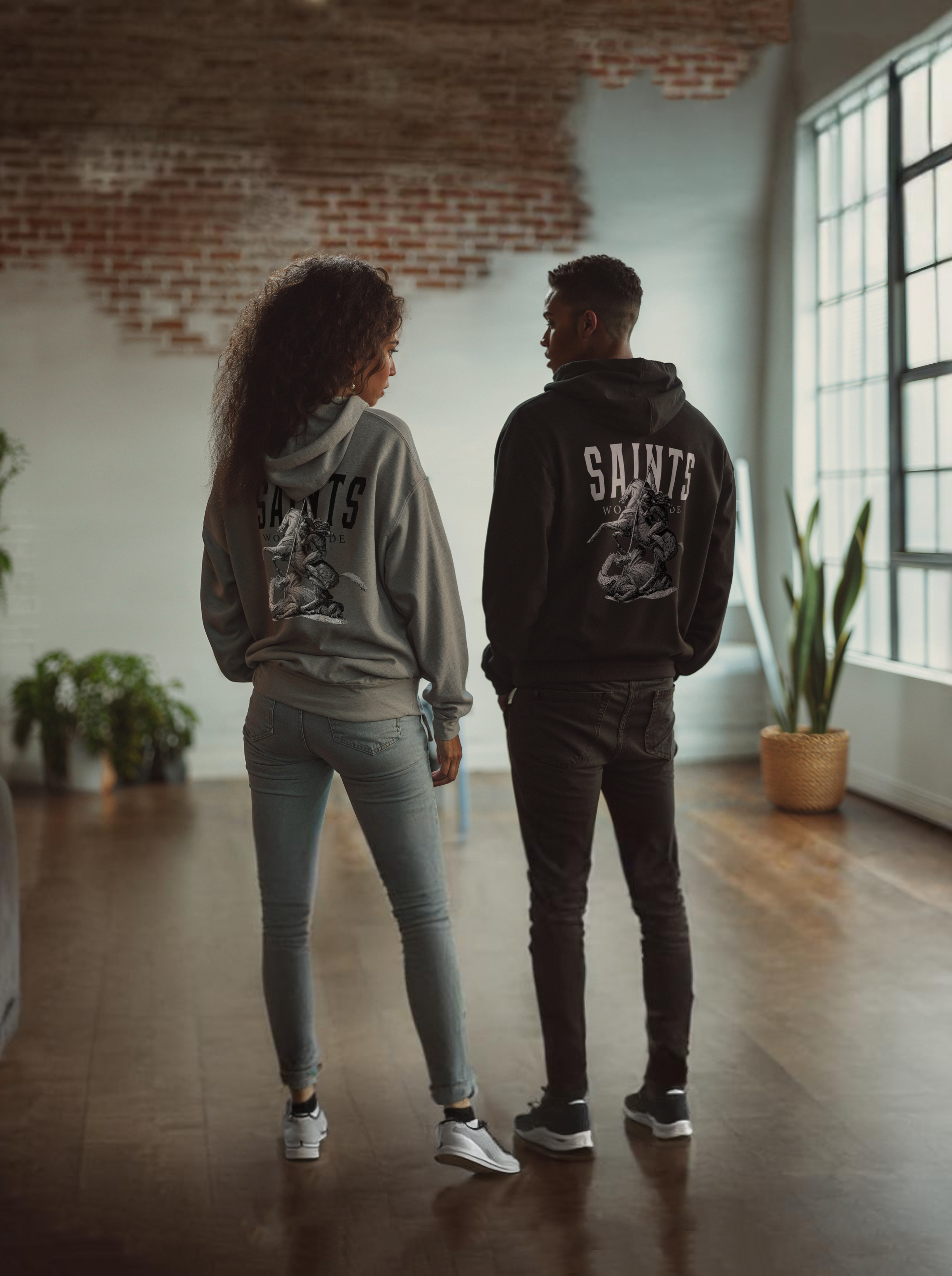 Man in a loft wearing black 'Saints Worldwide' hoodie looking at woman wearing grey  'Saints Worldwide' hoodie